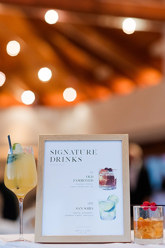 Signature drinks sign on a white tablecloth showing his and hers cocktails, framed with coupe and rocks glasses under warm bistro lights