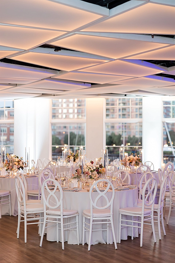 Reception tablescape with round tables, white linens, floral centerpieces, and tall glass hurricane candles by floor-to-ceiling city windows