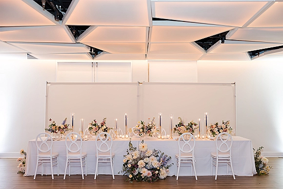 Head table decor with white tablecloth, low wedding sweetheart table decor florals, and pillar and taper candles against a white wall backdrop