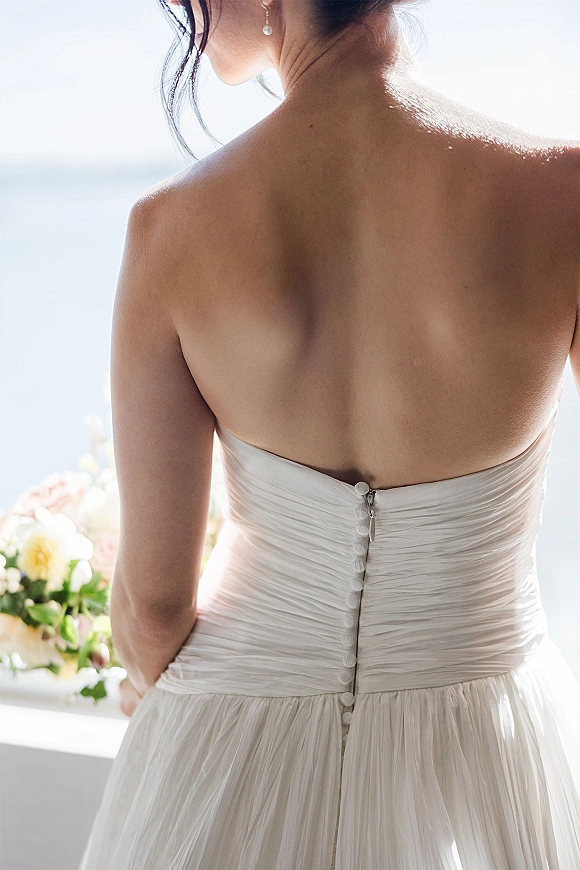 Wedding dress back in a strapless wedding dress with fabric-covered buttons, pearl drop earrings, and bouquet by a window with ocean view