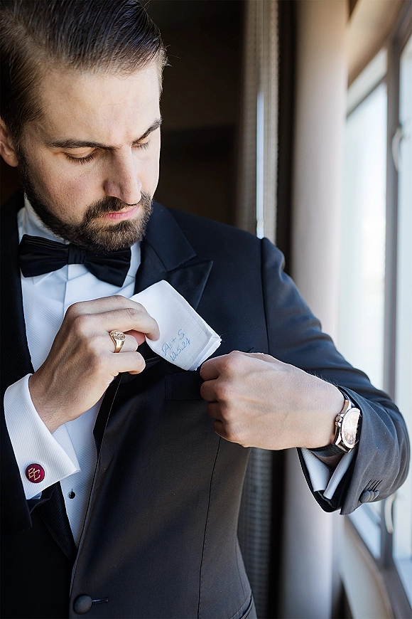 Groom portrait in a black tuxedo adjusting his pocket square by a window, showing bow tie, cufflinks, wristwatch, and signet ring