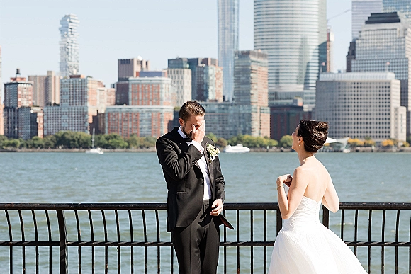 First look moment as bride approaches groom in black tuxedo by a waterfront railing with city skyline, boats, and blue sky behind