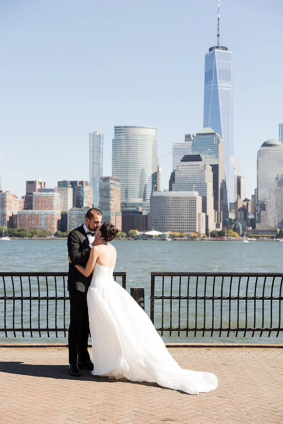 Wedding kiss portrait of bride and groom kissing by a waterfront railing, city skyline behind them under a clear blue sky, in tuxedo and dress
