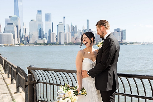 Couple portrait of bride and groom laughing by the waterfront, bride holding a white and green bouquet with city skyline behind them