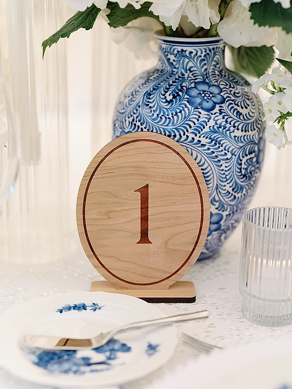 Wedding table number on a wood table number sign beside a blue and white ceramic vase with white flowers and a ribbed glass votive candle