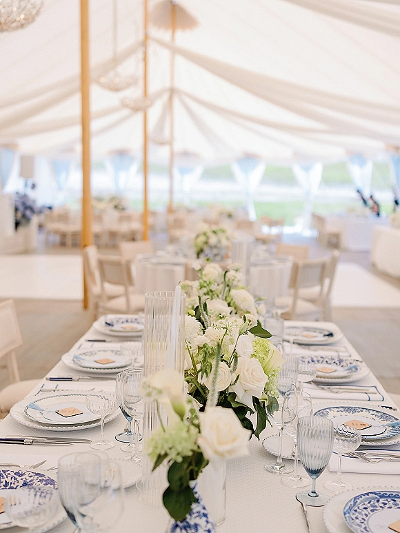 Reception tablescape with a wedding table centerpiece of white roses and greenery, blue patterned china, and glassware under a white tent