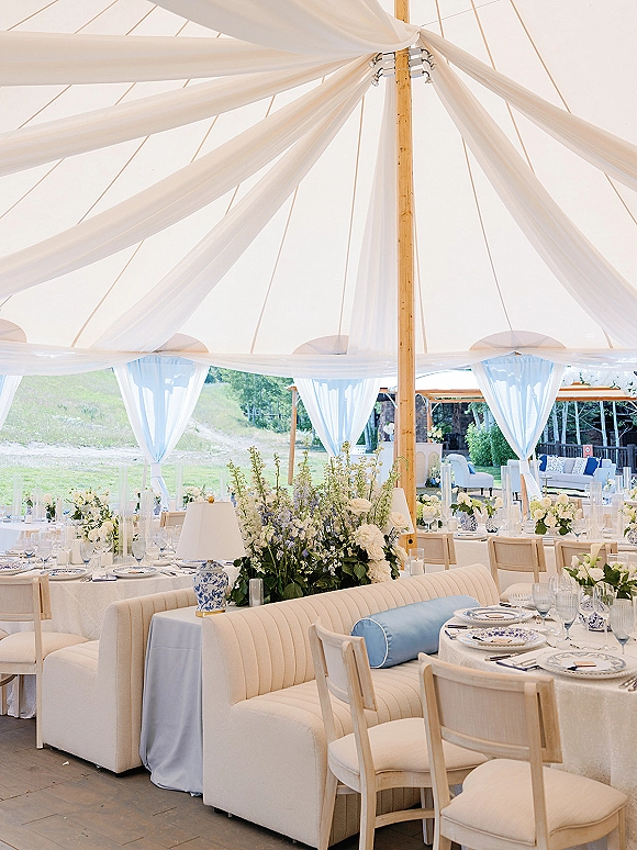 Reception tablescape in a tent wedding reception with round tables, blue and white china, lush florals, and a white draped ceiling over lawn and trees