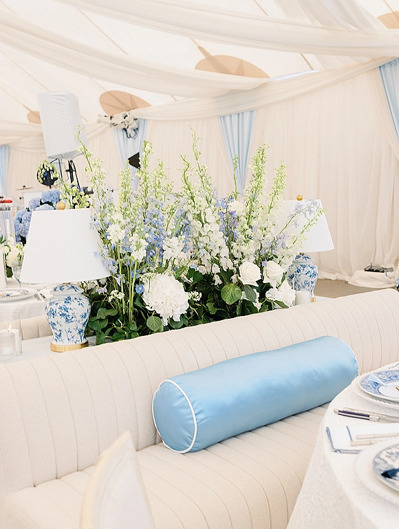 Reception lounge decor with a tufted banquette, chinoiserie lamps, and white-blue hydrangea and rose centerpiece under a draped tent ceiling