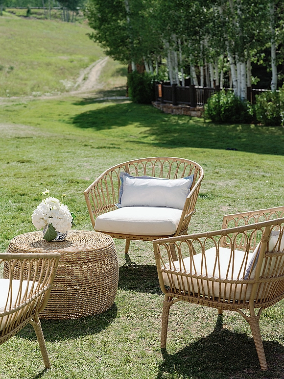 Outdoor lounge seating with rattan chairs, white cushions, and a round woven table topped with a blue-and-white vase of flowers on a hillside lawn