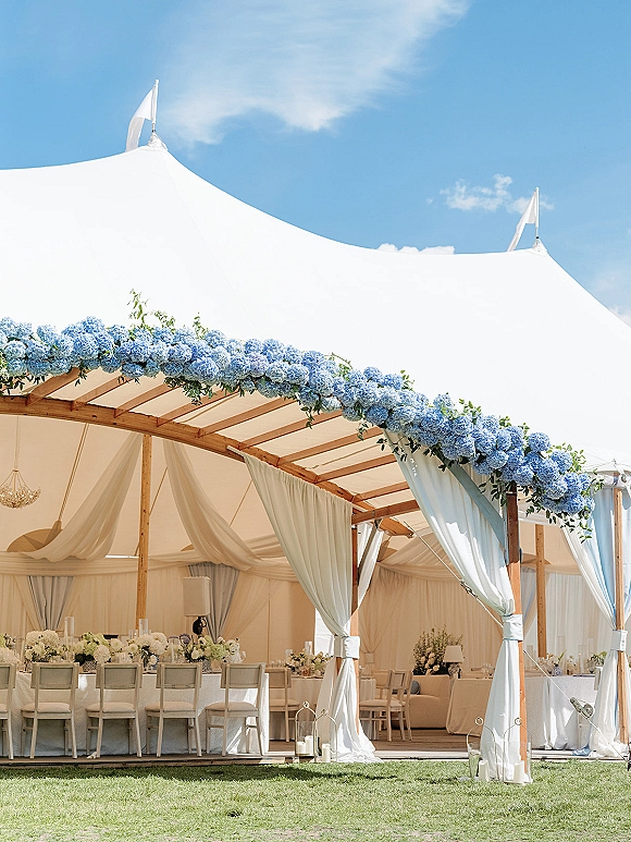 Wedding reception tent with outdoor tented reception draping, blue hydrangea garland, chandelier, and white tables on a grassy lawn under blue sky