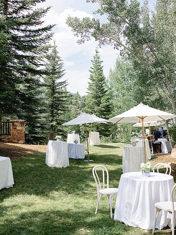 Cocktail hour setup with outdoor cocktail hour round tables in white linens and bistro chairs beneath patio umbrellas on a grassy lawn by pine trees