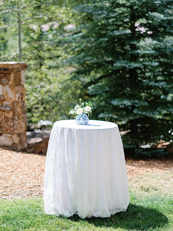 Wedding cocktail table with white lace tablecloth and blue-and-white vase of petite white flowers and greenery on a garden lawn by stone wall
