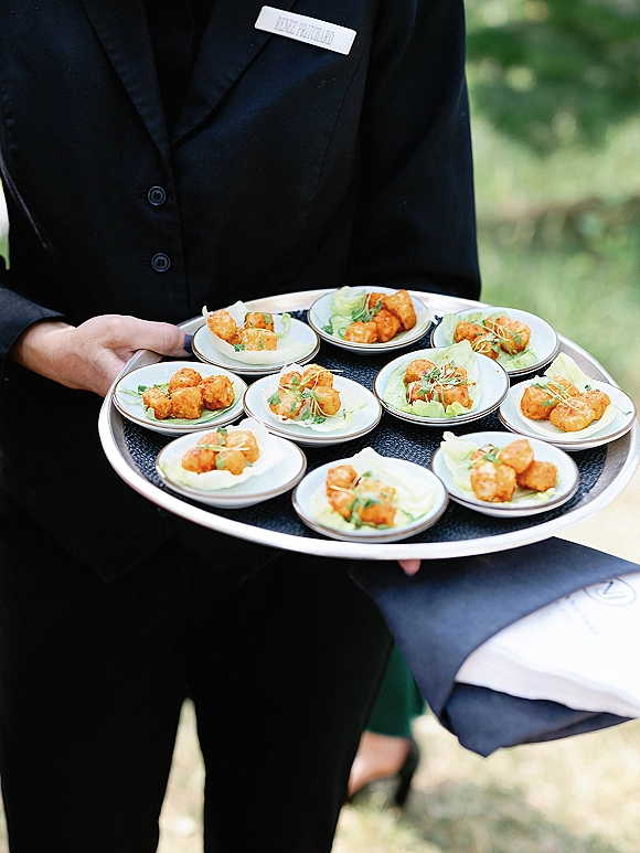 Wedding cocktail appetizers on passed hors d'oeuvres plates with lettuce garnish, held by a tuxedoed server on a tray outdoors on the lawn