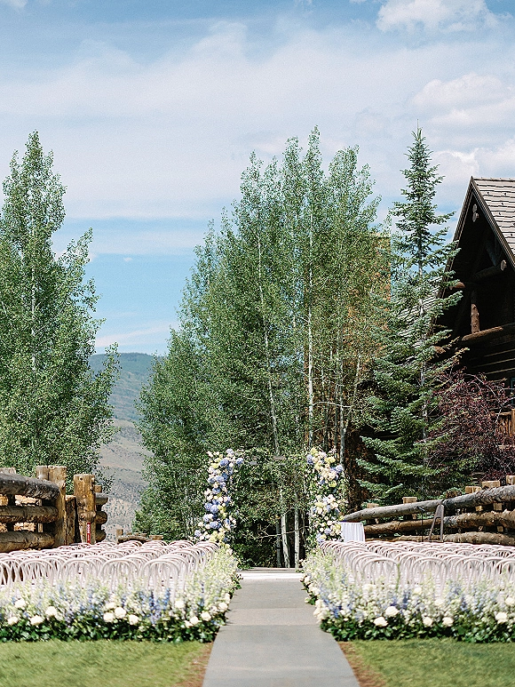 Ceremony aisle decor with an outdoor ceremony aisle lined by hydrangea markers and white chairs, facing a floral arch by a rustic lodge and mountains