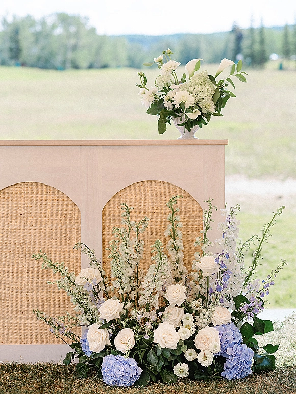 Wedding ceremony backdrop with arched panel and woven rattan, accented by blue hydrangeas and white roses in a grassy field