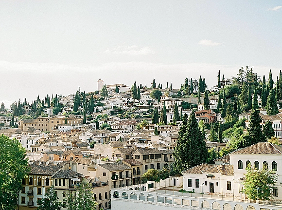 Hillside cityscape of a Mediterranean hillside town with whitewashed buildings, tiled rooftops and cypress trees under a blue sky