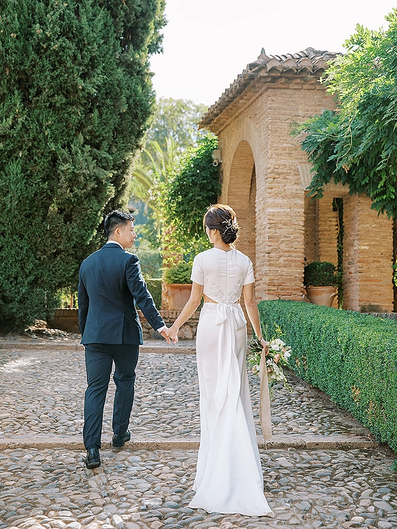 Couple portrait of bride and groom walking away holding hands, her bouquet with ribbon accent, along cobblestones under a stone archway