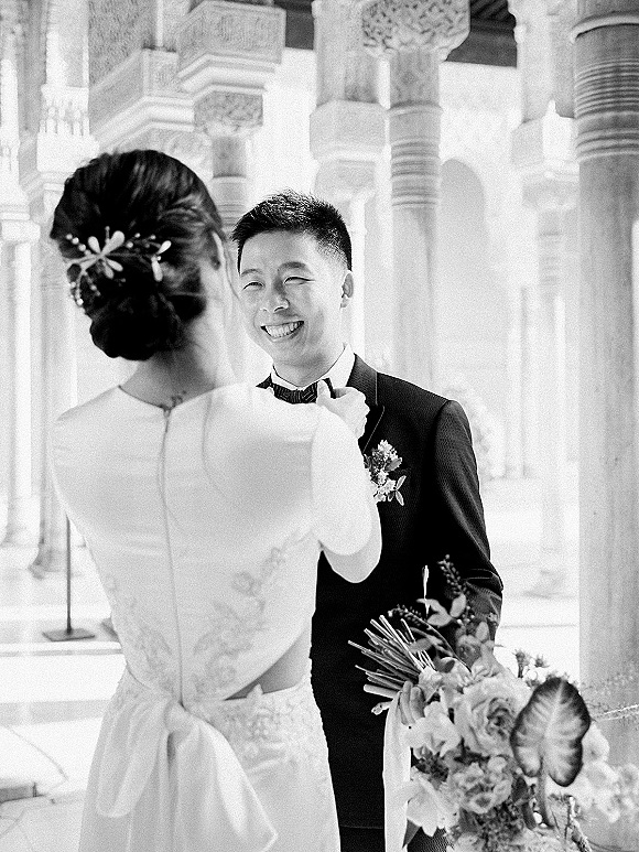First look moment as bride adjusts groom’s bow tie, face to face with bouquet and boutonniere beneath stone columns and arches.