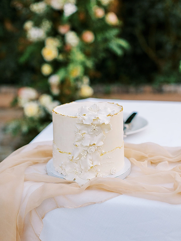 Wedding cake with white fondant and sugar flowers, gold leaf edging on a stand with chiffon runner, soft greenery bokeh behind
