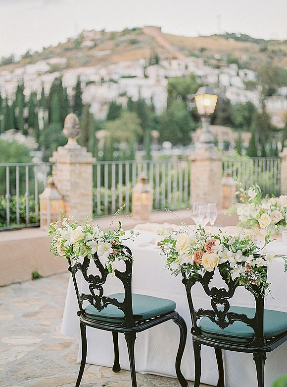 Reception table decor with white linen, lanterns and wine glasses, set with black chairs and floral garlands on a terrace overlooking a hillside town view