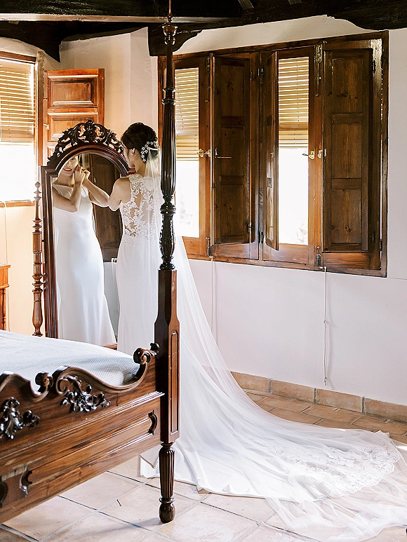 Bridal getting ready as the bride putting on earrings in an ornate mirror, wearing a lace-back gown with long veil in a bedroom