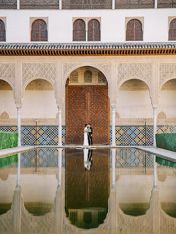 Couple portrait of bride in a gown holding a white bouquet and groom in a black suit, framed by an ornate courtyard archway and reflecting pool