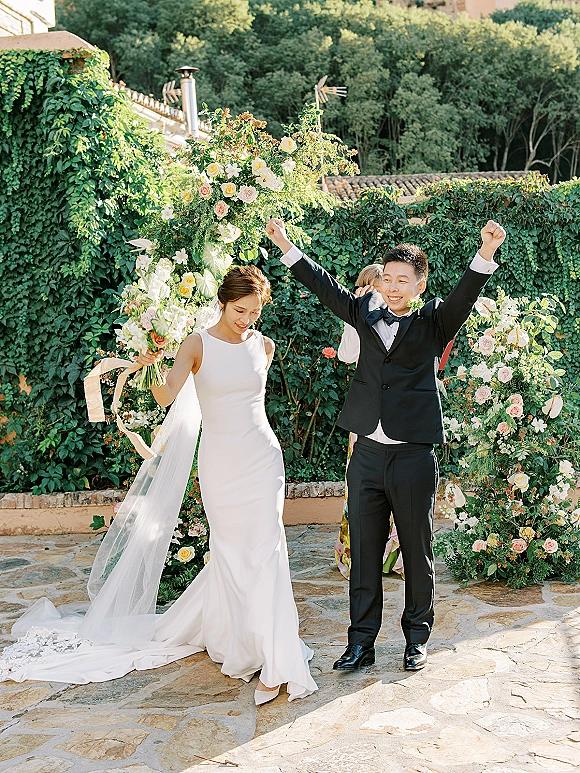 Recessional moment as bride and groom walk out cheering under a floral arch, bride holding bouquet with ribbon streamers in stone ivy courtyard