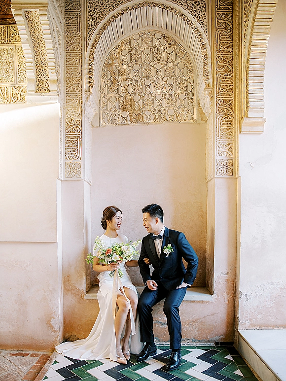 Couple portrait of bride and groom sitting and laughing, bride holding bouquet beside groom in tuxedo before an ornate arched niche backdrop