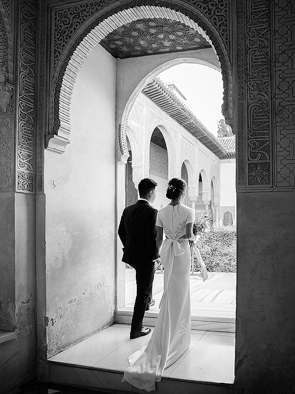 Couple portrait in a black and white wedding portrait, bride and groom walking away under an ornate archway, bouquet in hand