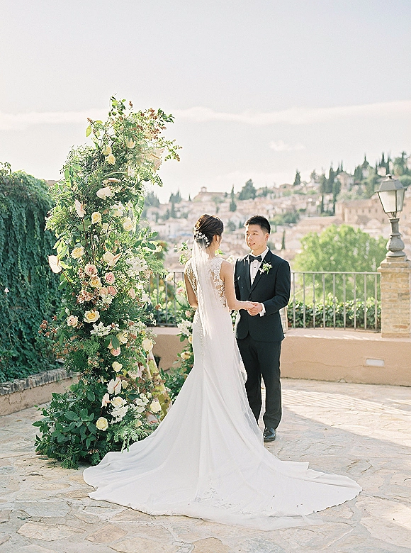 Ceremony moment as bride and groom hold hands beneath a floral arch of roses and greenery on a stone terrace overlooking a hillside town