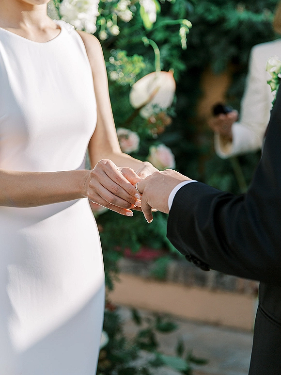 Ring exchange as the groom places a wedding band on the bride’s hand, with anthurium and rose greenery at an outdoor garden ceremony
