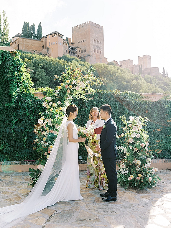 Wedding vows as bride and groom face the officiant at an outdoor wedding ceremony on a stone patio beside an ivy-covered castle wall