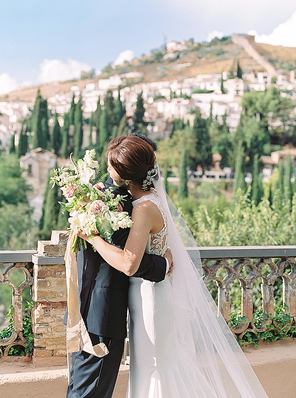 Couple portrait of bride and groom hug on a stone terrace, bride holding bouquet and veil trailing, hillside village and cypress trees behind