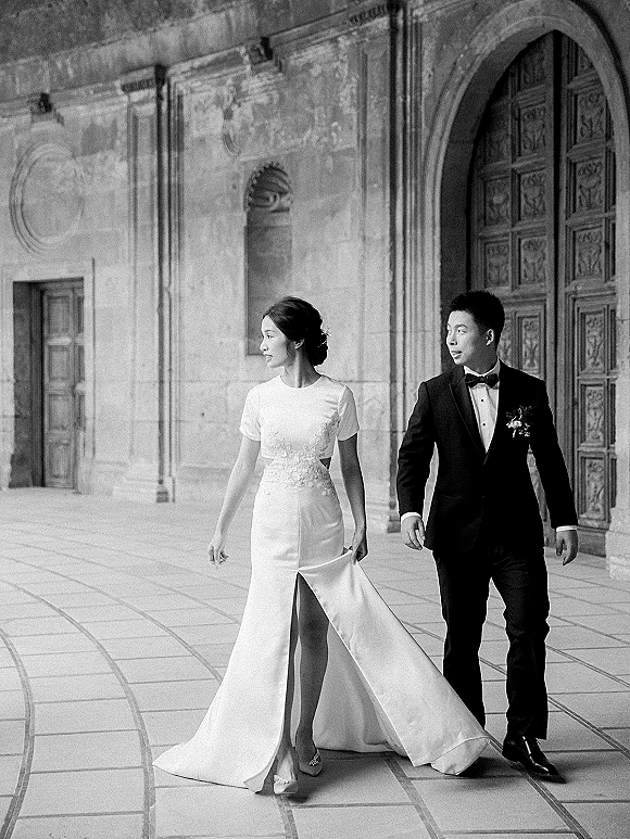 Couple portrait of bride and groom walking side by side, bride holding dress train, in a stone courtyard with arched doorway