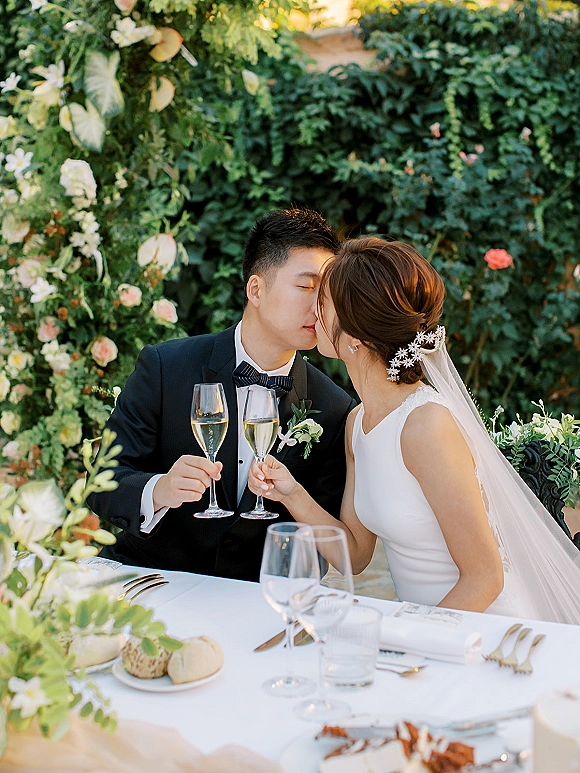 Wedding kiss portrait of bride and groom with champagne flutes, her veil draped at a sweetheart table against a lush greenery wall