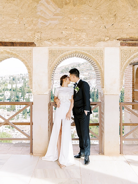 Wedding kiss portrait of bride and groom kiss beneath a carved stone archway on a balcony overlooking the city and hills