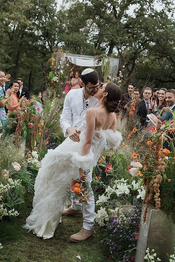 Wedding kiss as bride in ruffle dress and groom in white suit dip in floral-lined aisle under arch, guests cheering in garden greenery