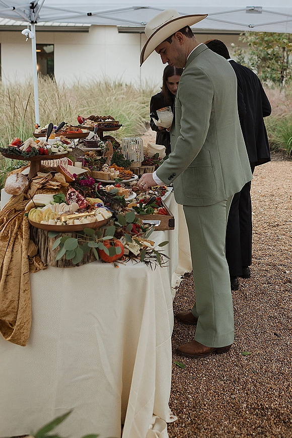 Wedding grazing table with a charcuterie spread of cheeses, fruit, bread, and trays on wooden risers under a white tent with eucalyptus accents