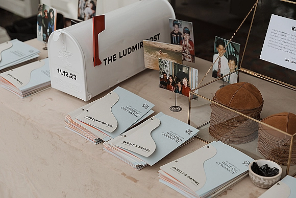Wedding program fans in light blue and white stacked on an indoor tabletop with RSVP cards, mailbox holder, photo prints, and gold frame