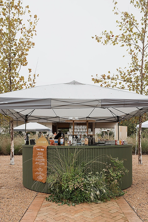 Wedding bar setup under a canopy tent with cocktail menu sign, liquor bottles and glassware, plus greenery accents in a garden setting