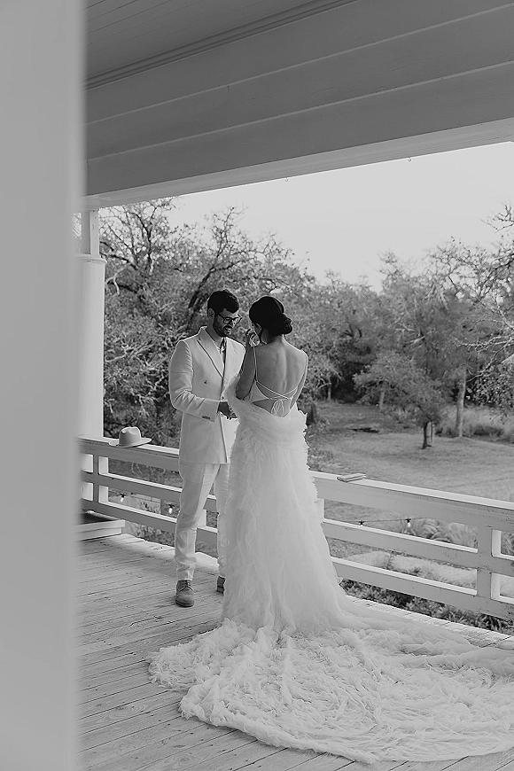 Wedding couple portrait in black and white holding hands on a rustic porch, bride’s backless gown with long train under string lights