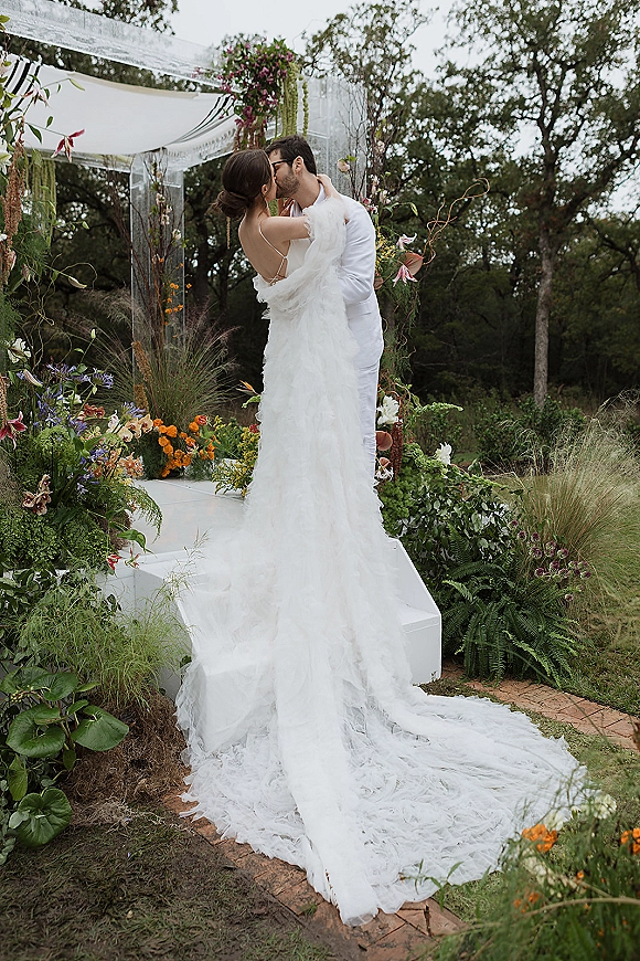 Wedding kiss portrait of bride and groom kissing beneath a fabric canopy arch, her backless gown’s long train flowing over grass