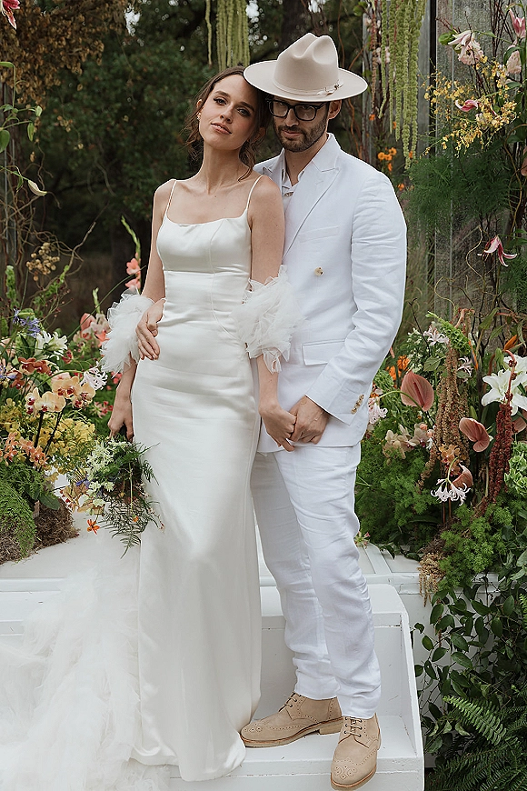 Couple portrait of bride and groom embrace, he in a white suit and cowboy hat on white steps beneath hanging greenery outdoors