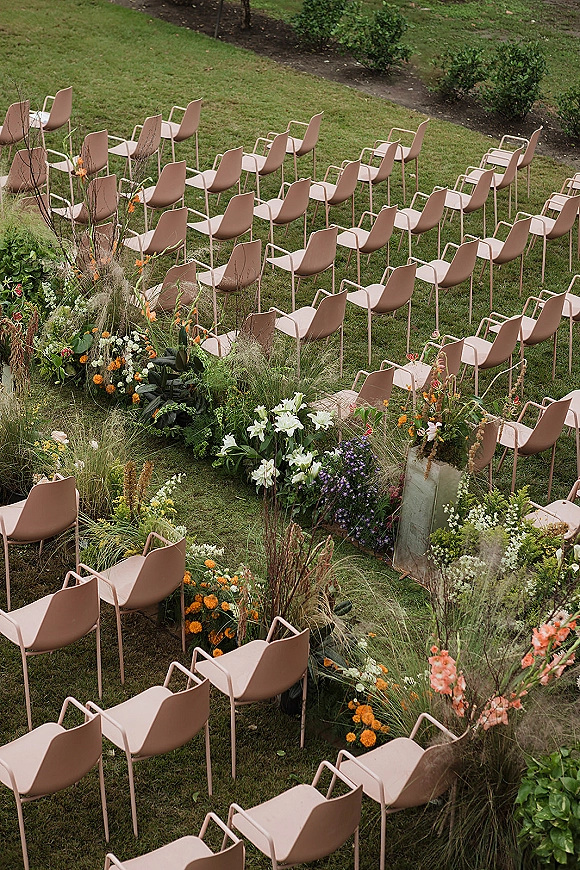 Outdoor ceremony setup with garden ceremony seating along a curved aisle, lined with meadow-style florals and greenery on a lawn