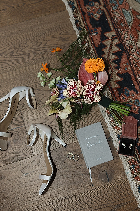 Wedding detail flat lay with bridal accessories flat lay, heels, orchid bouquet, rings, vow book and jewelry on wood floor with vintage rug