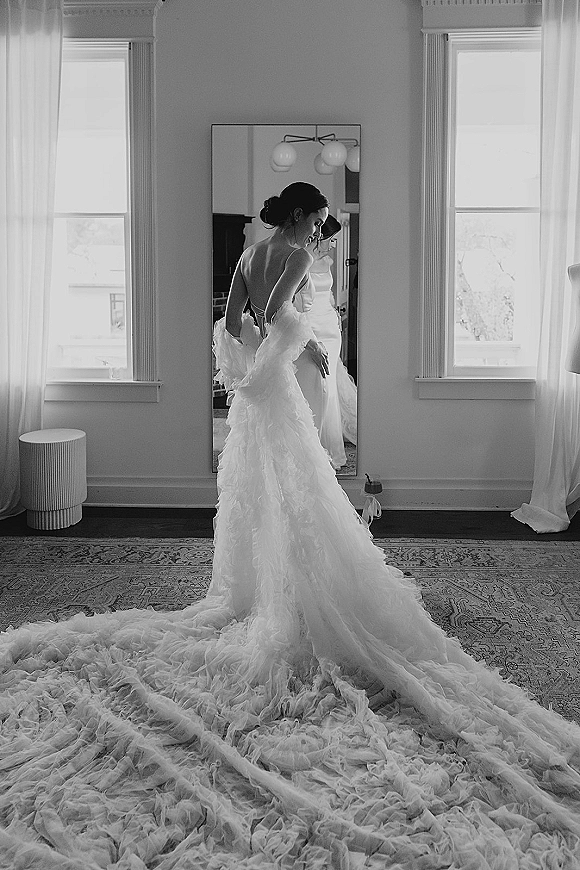Bridal portrait of a bride in a black and white bridal portrait, wearing an open-back gown with ruffled tulle train by tall windows