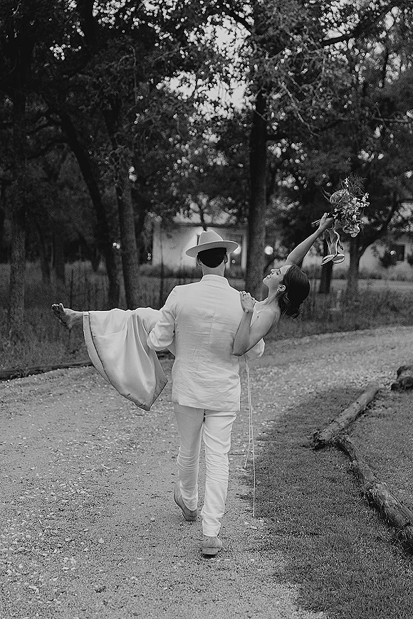 Couple portrait of groom carrying bride as she raises a bridal bouquet with ribbon streamers along a tree-lined path in a park