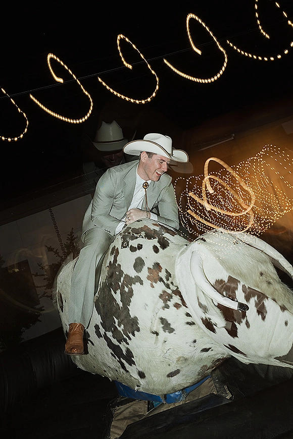 Mechanical bull ride at a wedding reception with groom in cowboy hat, light gray suit and bolo tie under string lights indoors