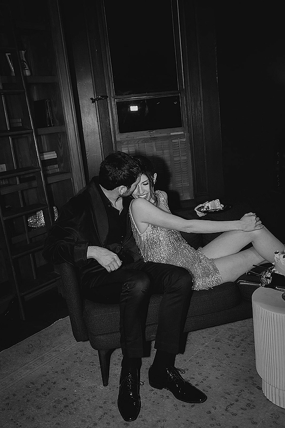 Couple portrait in a wood-paneled library lounge, groom kissing bride’s cheek as she sits in a sequin mini dress with cake and champagne