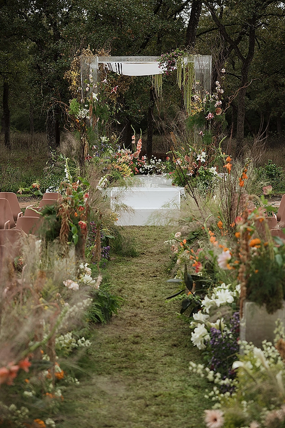 Ceremony aisle decor with wildflower wedding aisle blooms lining the grass, leading to a wood arch with white drape and hanging greenery in a forest setting